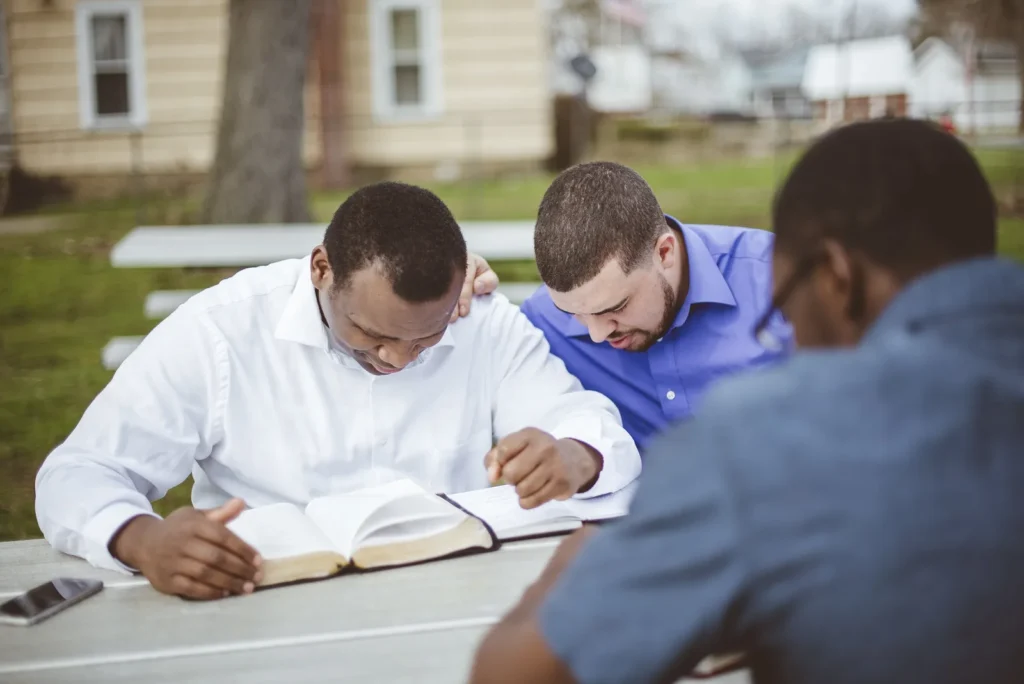 Tres hombres leen juntos la Biblia en una mesa al aire libre; uno apoya la mano en el hombro de otro