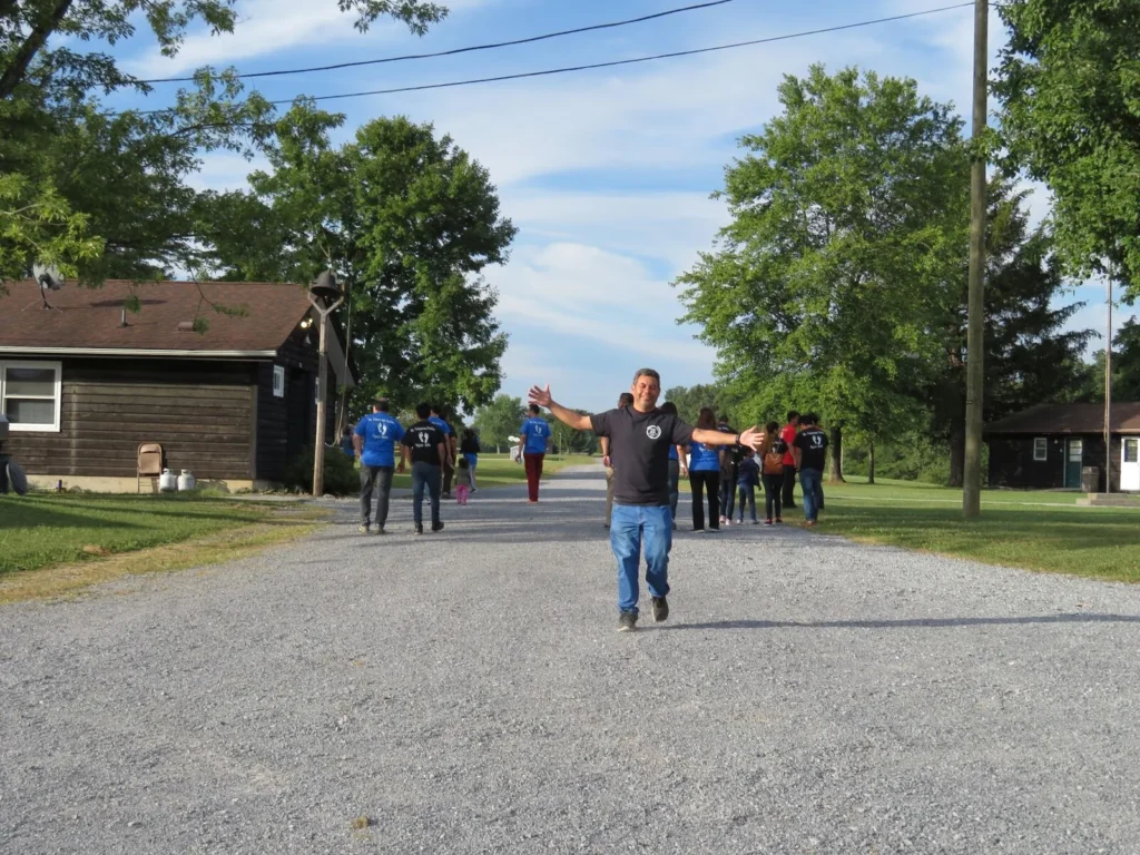 Un hombre abre los brazos en señal de bienvenida en el camino de grava del campamento mientras la congregación camina detrás