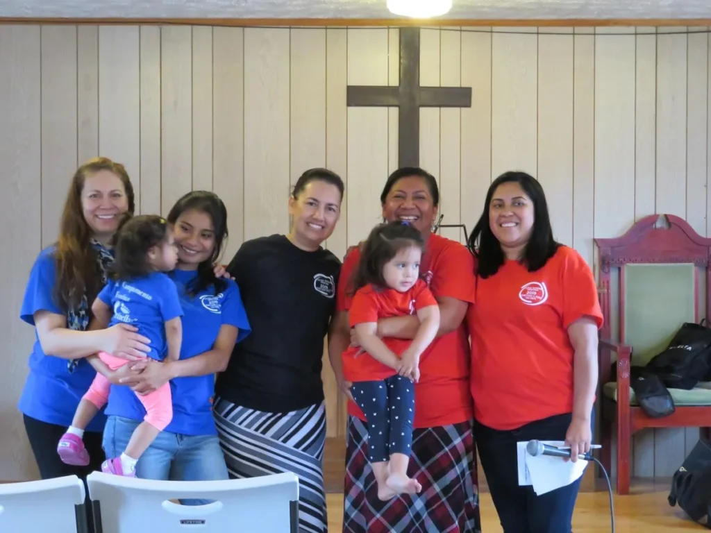Mujeres de la iglesia con camisetas del campamento sonríen junto a la cruz, dos de ellas cargando a niñas pequeñas