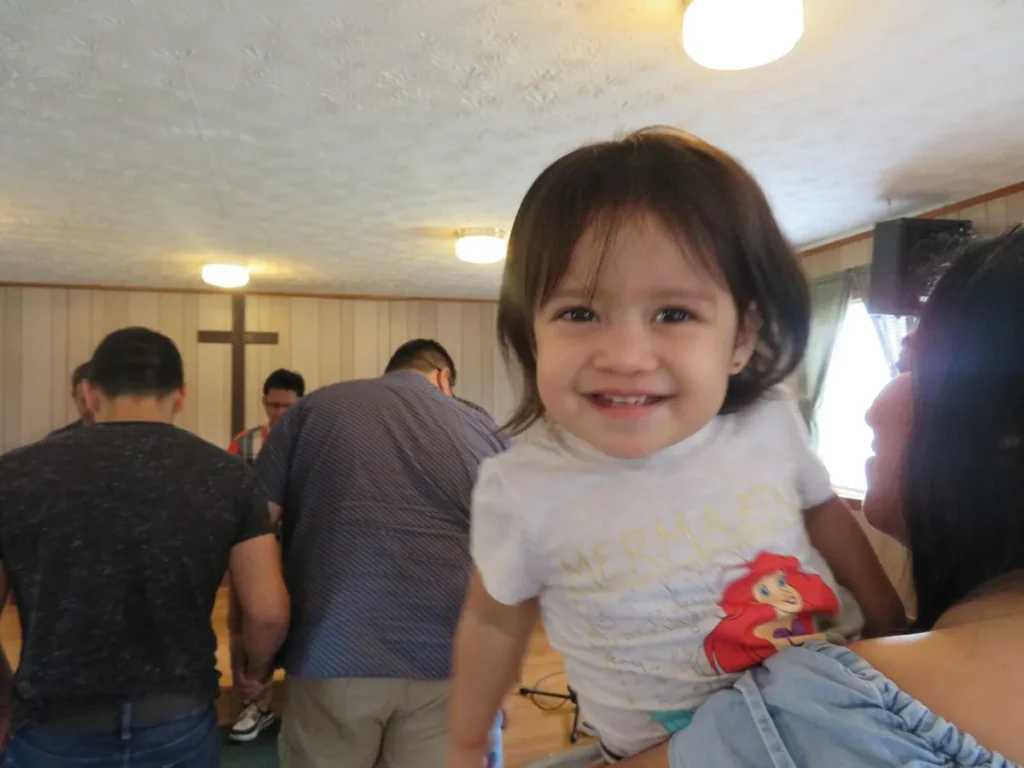 Una niña pequeña sonríe en brazos de su mamá mientras los hermanos oran al fondo
